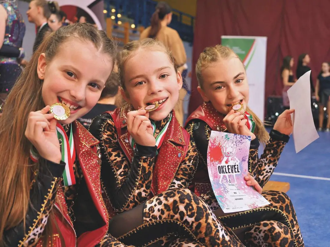 Három kislány nevetve, éremmel a szájukban és oklevéllel egy verseny után